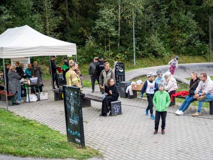 Wydarzenie plenerowe obok toru pumptrack. Po lewej stoi biały namiot ze stanowiskiem rejestracji, wokół zebrali się dorośli i dzieci. Na pierwszym planie chłopiec w zielonej kurtce, w tle leśny krajobraz.