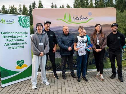 Chłopiec z numerem startowym 208 trzyma dyplom na tle ścianki z logo Istebnej, otoczony dorosłymi i sędziami. Po lewej stronie stoi baner Gminnej Komisji Rozwiązywania Problemów Alkoholowych w Istebnej. Uczestnicy patrzą w stronę aparatu.