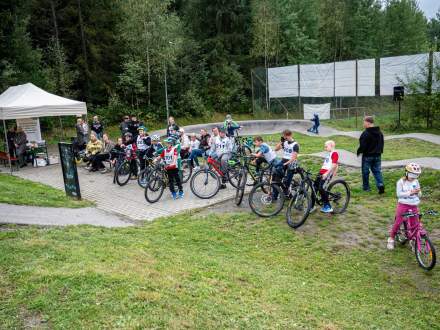 Szeroki kadr na plenerowe zawody rowerowe na torze pumptrack. Dzieci w kaskach z numerami startowymi usytuowane są z rowerami na starcie. Po lewej znajduje się biały namiot, a w tle gęsty, zielony las.