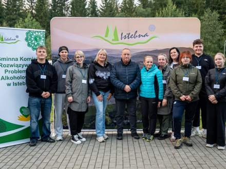 Grupa organizatorów i wolontariuszy stoi razem do pamiątkowego zdjęcia na tle ścianki z logo Istebnej. Po lewej stronie znajduje się baner profilaktyczny. Wszyscy patrzą w obiektyw aparatu.