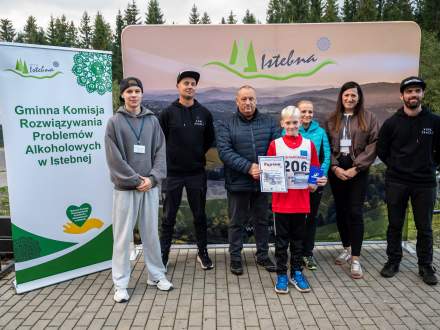 Chłopiec z numerem 206 trzyma dyplom, stojąc z grupą dorosłych i sędziami na tle ścianki sponsorskiej Istebnej. Po lewej zielony baner, w tle las. Wszyscy patrzą w obiektyw
