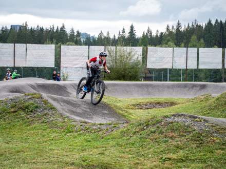 Młody rowerzysta w kasku i sportowym stroju z numerem startowym pokonuje wzniesienie na torze pumptrack. W tle widać profilowaną trasę, białe ekrany ogrodzenia oraz gęsty, las świerkowy.