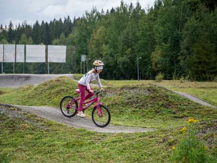 Dziecko w kasku i jasnej bluzie jedzie na różowym rowerze po asfaltowym torze pumptrack. W tle widać trawiaste pagórki i gęsty las pod zachmurzonym niebem.