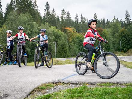 Czworo dzieci w kaskach i z numerami startowymi jedzie na rowerach górskich po torze pumptrack. Jeden chłopiec jedzie z przodu po prawej stronie, a troje pozostałych podąża za nim po lewej. W tle rośnie las.