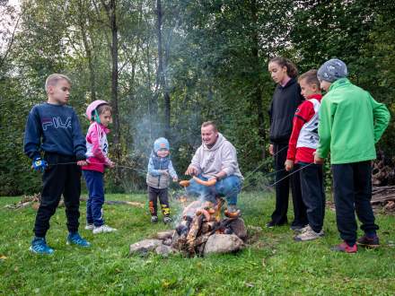 Mężczyzna i grupa dzieci pieką kiełbaski na kijach nad ogniskiem rozpalonym na trawie. Uczestnicy stoją w kręgu w otoczeniu zielonych drzew i krzewów.