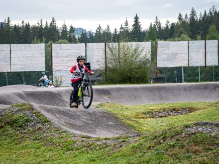 Chłopiec w kasku i z numerem startowym 203 jedzie na rowerze górskim po asfaltowym torze pumptrack. W tle widać ogrodzenie oraz gęsty, zielony las.