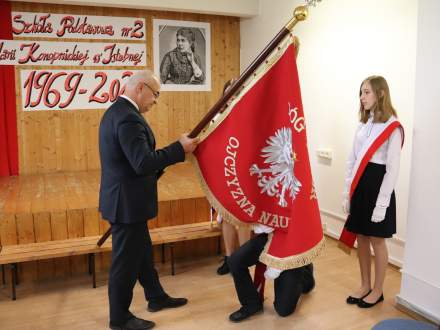 Dyrektor Szkoły Podstawowej nr 2 Bogdan Ligocki przekazuje sztandar Szkolnemu Pocztowi Sztandarowemu; sztandarowy przyklęka by go ucałować; sztandar skierowany orłem białym na czerwonym tle do publiczności
