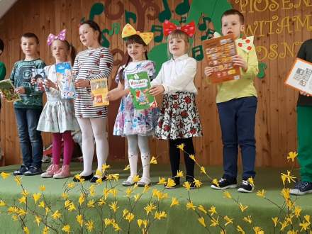 Uczniowie pierwszej klasy na scenie – 4 chłopców i 4 dziewczynki; zielona scena, przed sceną żółte, wiosenne kwiaty. Dzieci maja w dłoniach, kolorowe książeczki z bajkami
